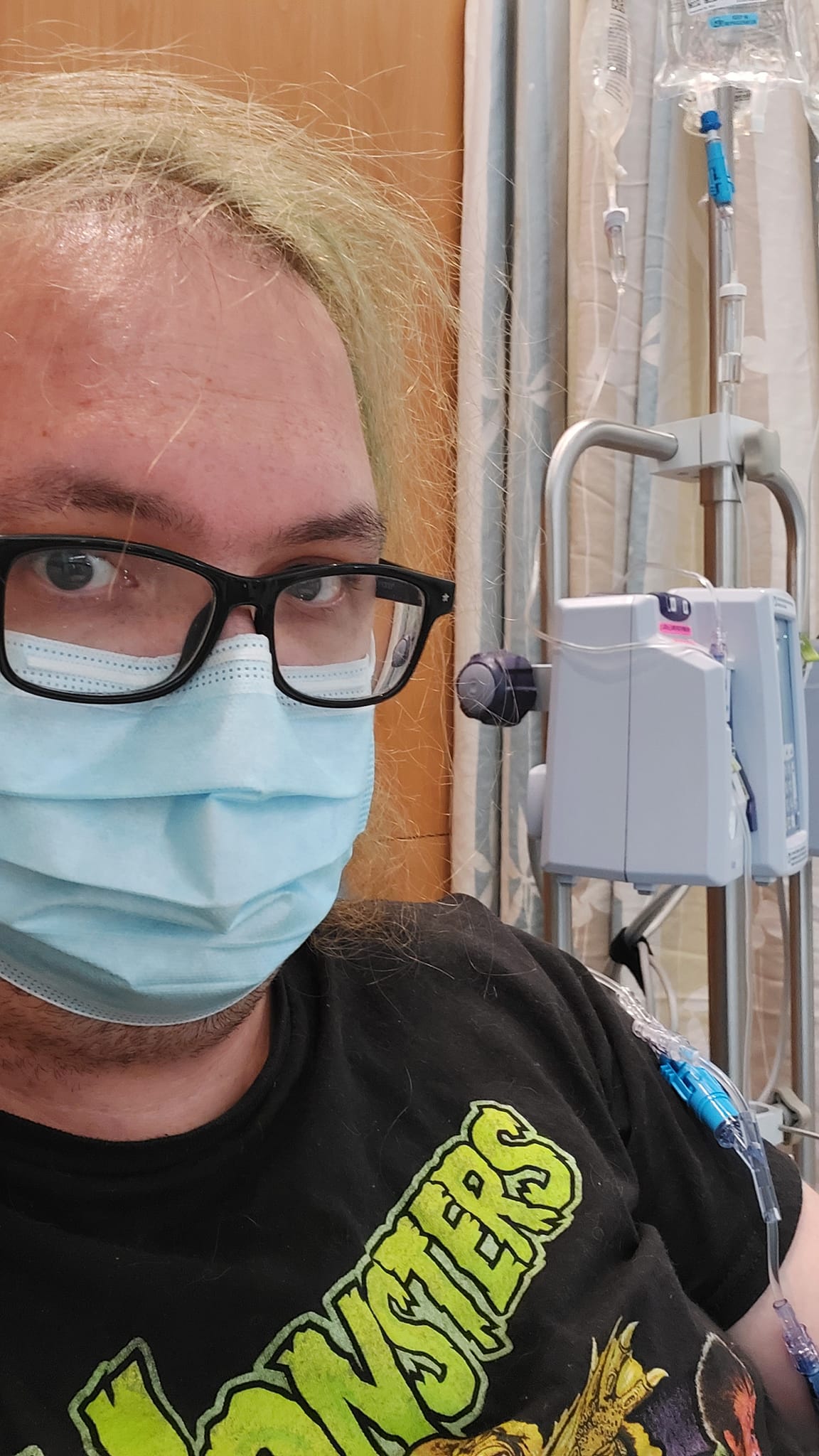 A man with green hair wearing a black Universal Monsters shirt sits in a hospital room, IV bags and pumps visible in the background